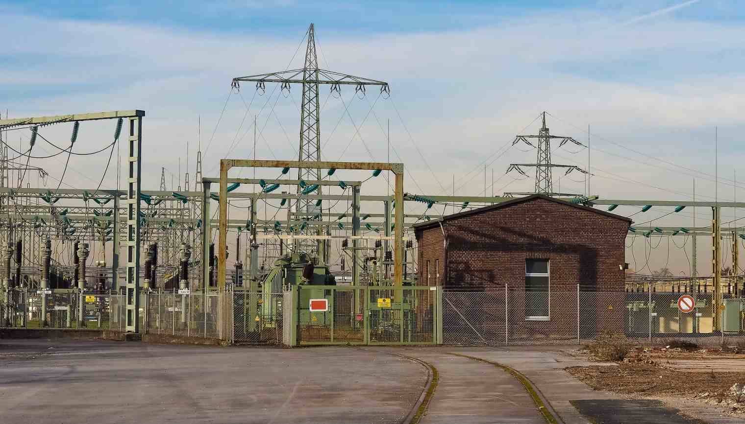A high-voltage substation behind a security fence, with transmission towers beyond